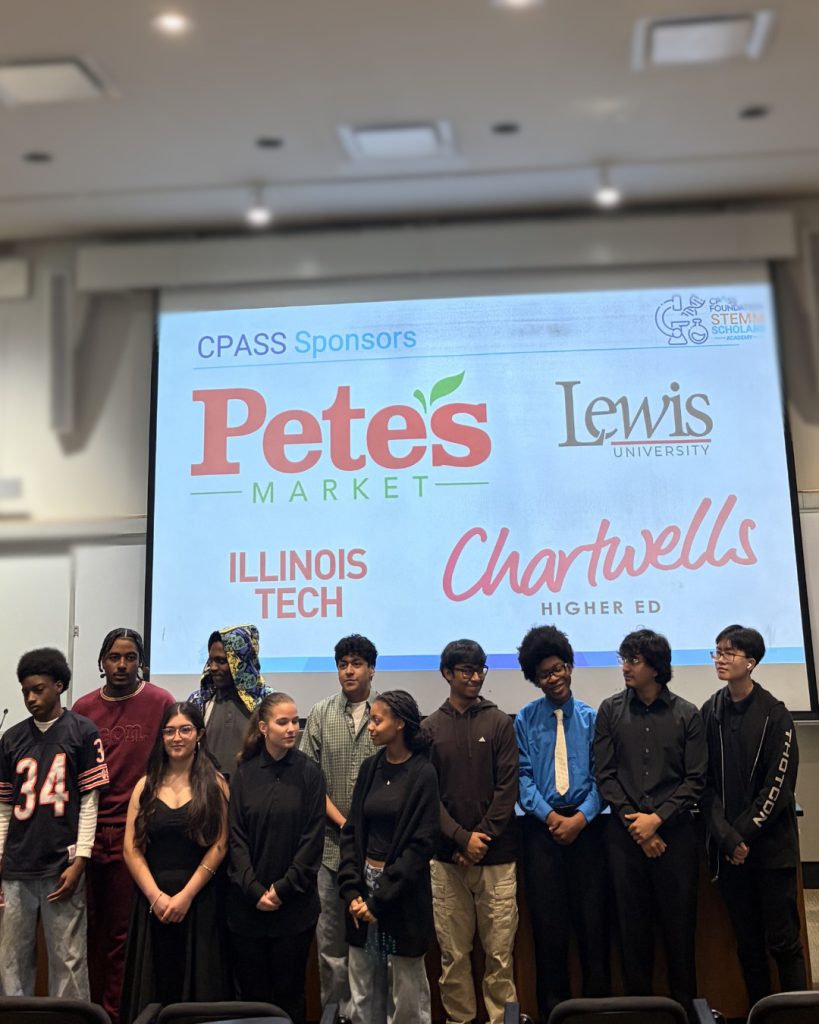 Group of students standing in a line at the front of a lecture hall beneath a large projected slide labeled “CPASS Sponsors.” The screen displays logos for Pete’s Market, Lewis University, Illinois Tech, and Chartwells Higher Ed. The classroom includes a wide projection screen, overhead lighting, whiteboard panels, and tiered seating in the foreground.